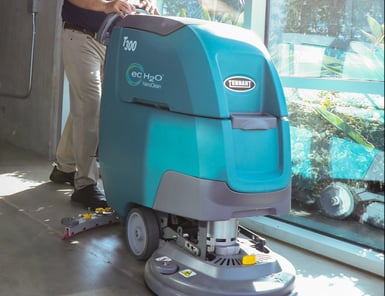 A khaki-clad worker operates a Tennant ec-H2O commercial floor scrubber to clean an interior tile floor near a glass storefront.