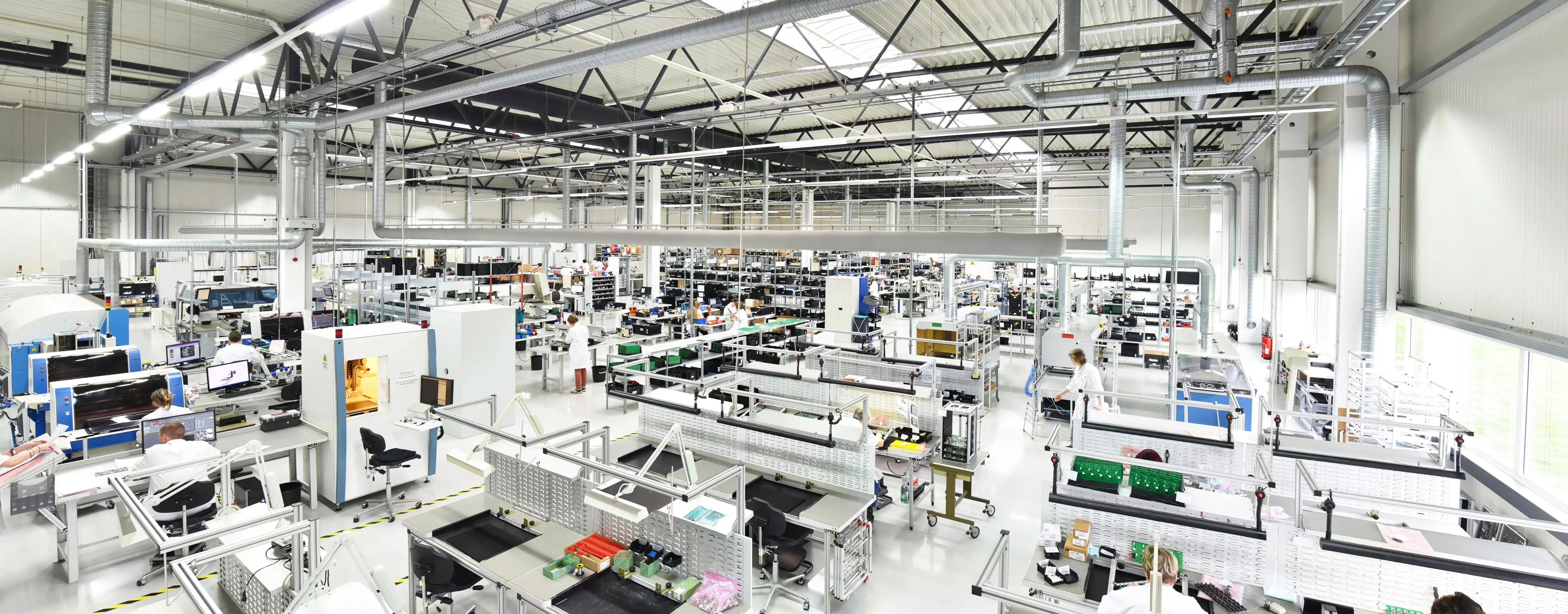Wide interior view of a large manufacturing floor with workers in white lab coats at assembly stations under overhead ductwork and skylights.