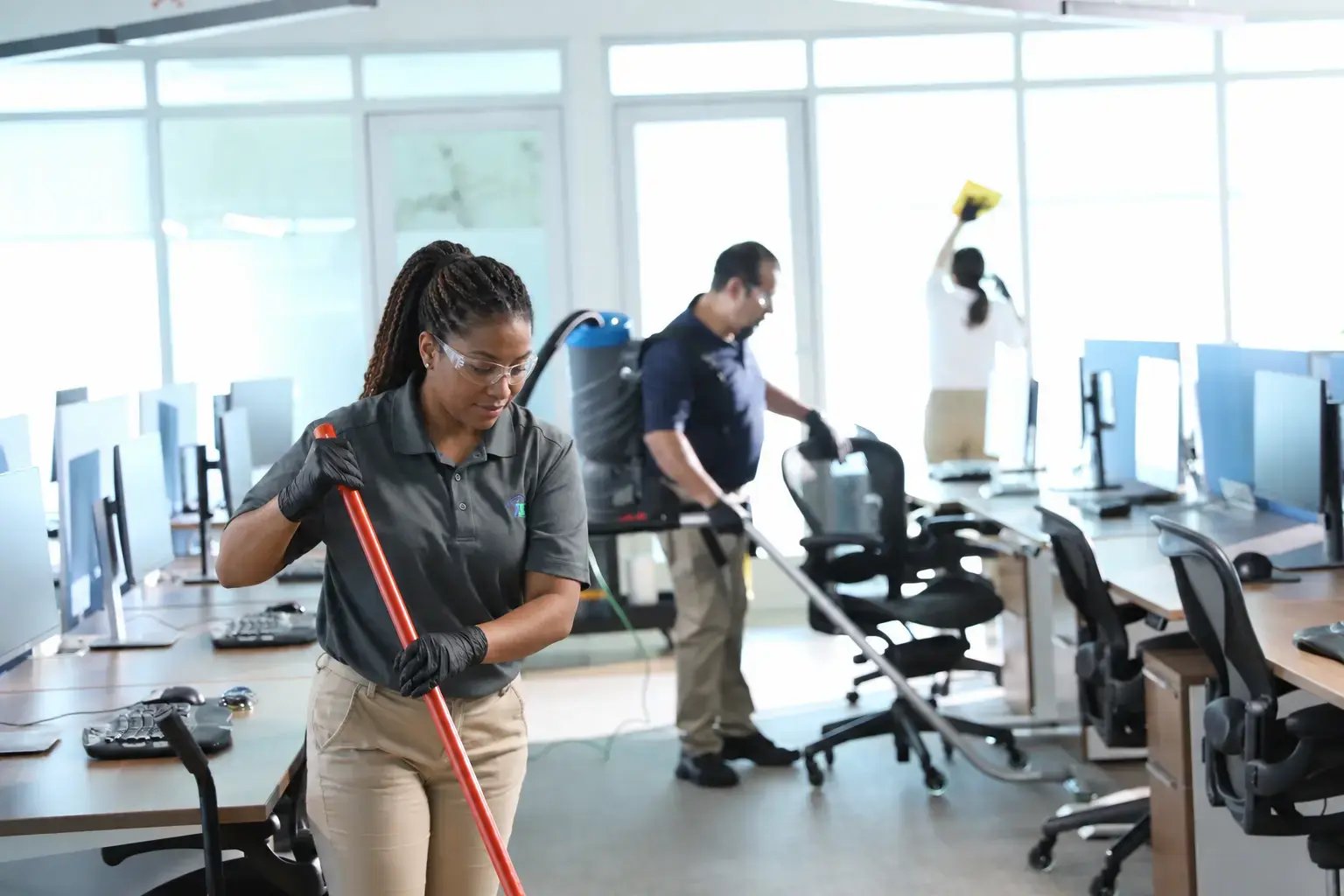 A cleaning technician sweeps the floor with a broom in a bright computer lab while two coworkers clean workstations and windows in the background.