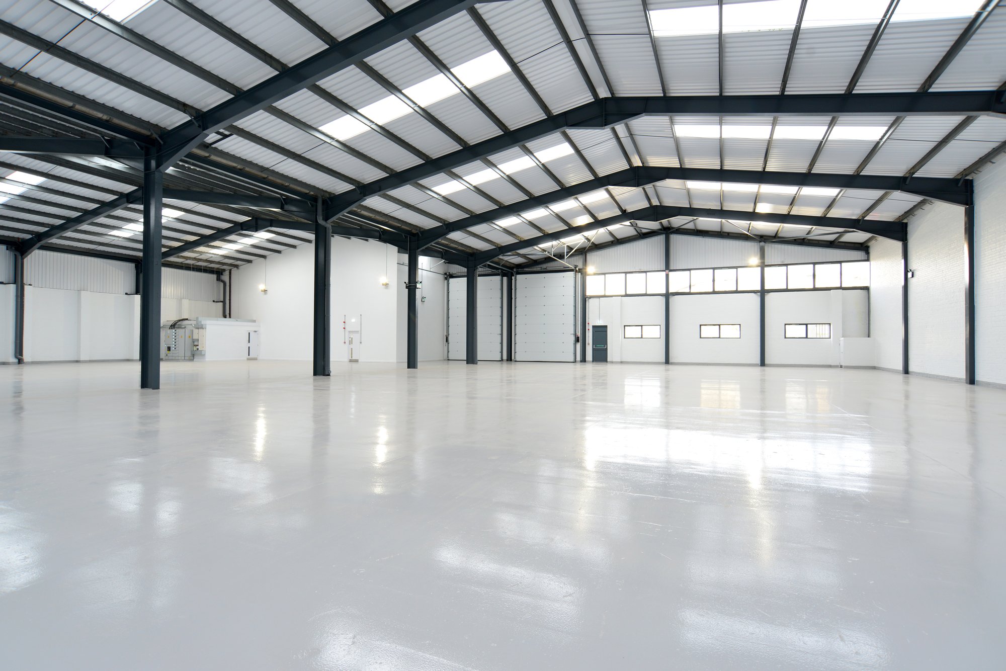 Clean, empty warehouse interior with polished white epoxy floors, steel beam ceiling with skylights, and large roll-up loading doors.Clean, empty warehouse interior with polished white epoxy floors, steel beam ceiling with skylights, and large roll-up loading doors.