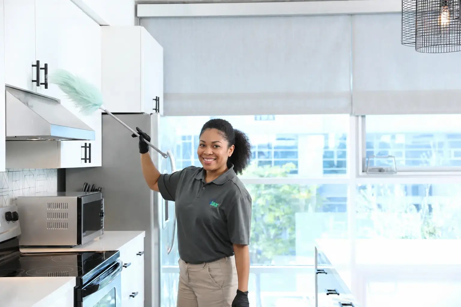 An iNX cleaning technician in uniform smiling while dusting kitchen cabinets with an extendable duster in a modern office break room.