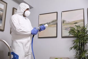 A cleaning technician in a hazmat suit sprays disinfectant on framed artwork in a lobby.