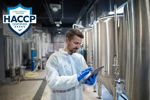 A HACCP Certified badge overlaid on a photo of a technologist in a white protective coat and blue gloves reviewing a tablet inside a food and beverage manufacturing facility.