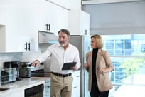 Two people in a modern kitchen, one holding a tablet and pointing at an appliance while discussing it with the other.