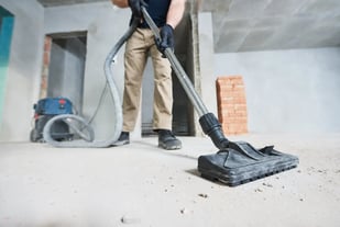 A cleaning technician uses an industrial vacuum to clear dust and debris from a bare concrete floor in an unfinished construction space.