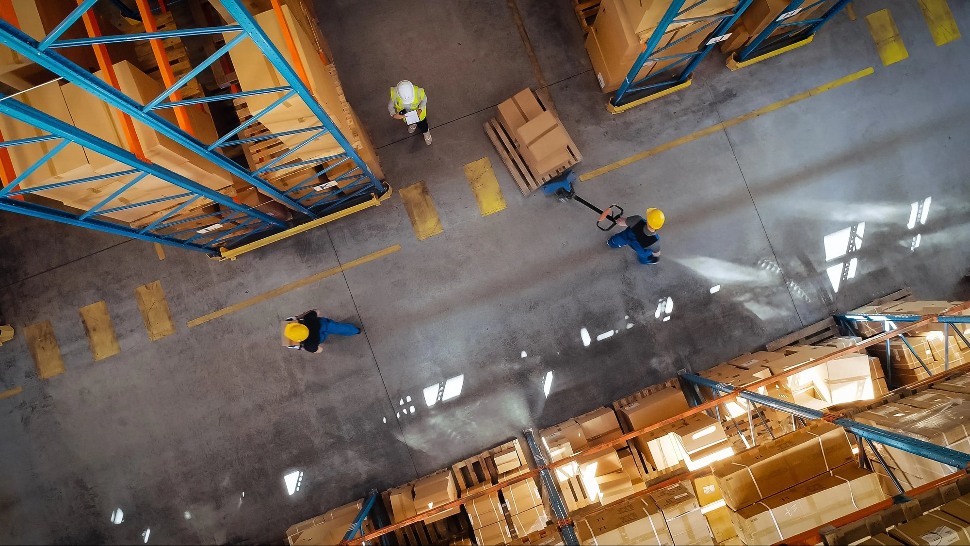 Aerial view of three warehouse workers in hard hats and safety vests moving pallets with a hand truck between tall storage racks stocked with cardboard boxes.