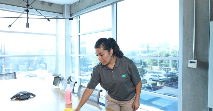 An iNX day porter in a branded uniform wiping down a conference room table in a corporate office setting.